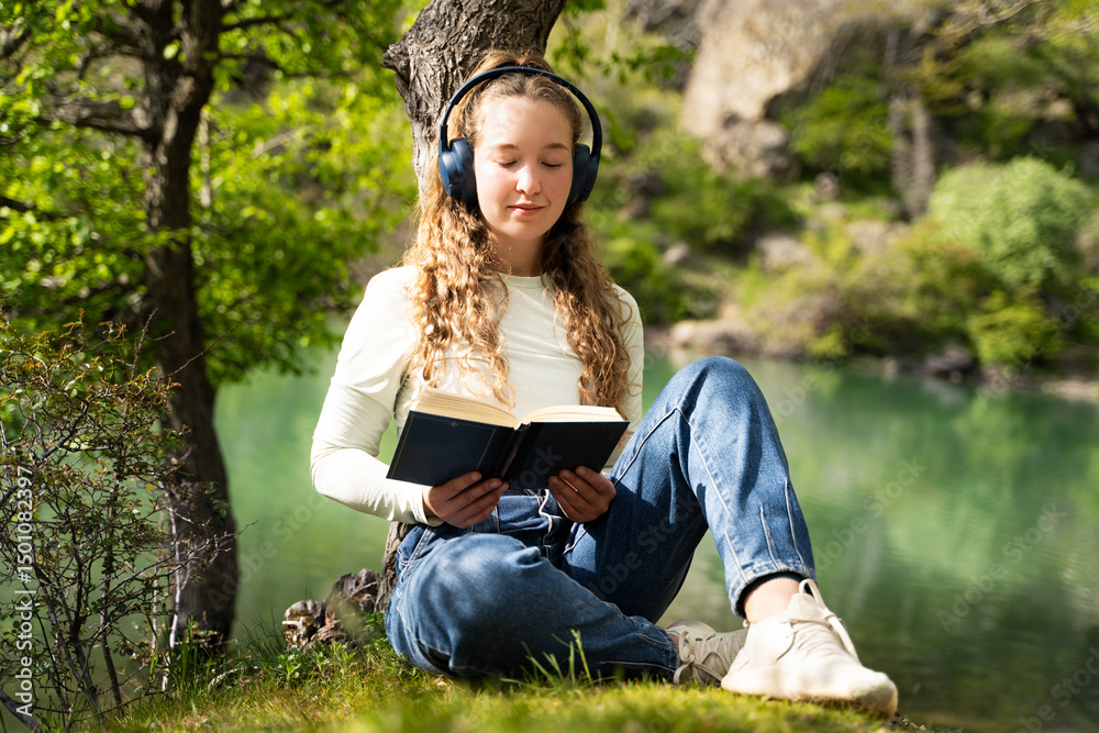 Young woman enjoying music and reading book in nature relaxing by the lake wearing headphones, peaceful outdoor lifestyle, mindfulness and digital detox, spring forest background, front view full body Young woman enjoying music and reading book in nature relaxing by the lake wearing headphones, peaceful outdoor lifestyle, mindfulness and digital detox, spring forest background, front view full body