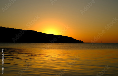 Tranquil seascape at sunset with golden reflections on the calm water and the sun setting behind a silhouette of a coastal hill.