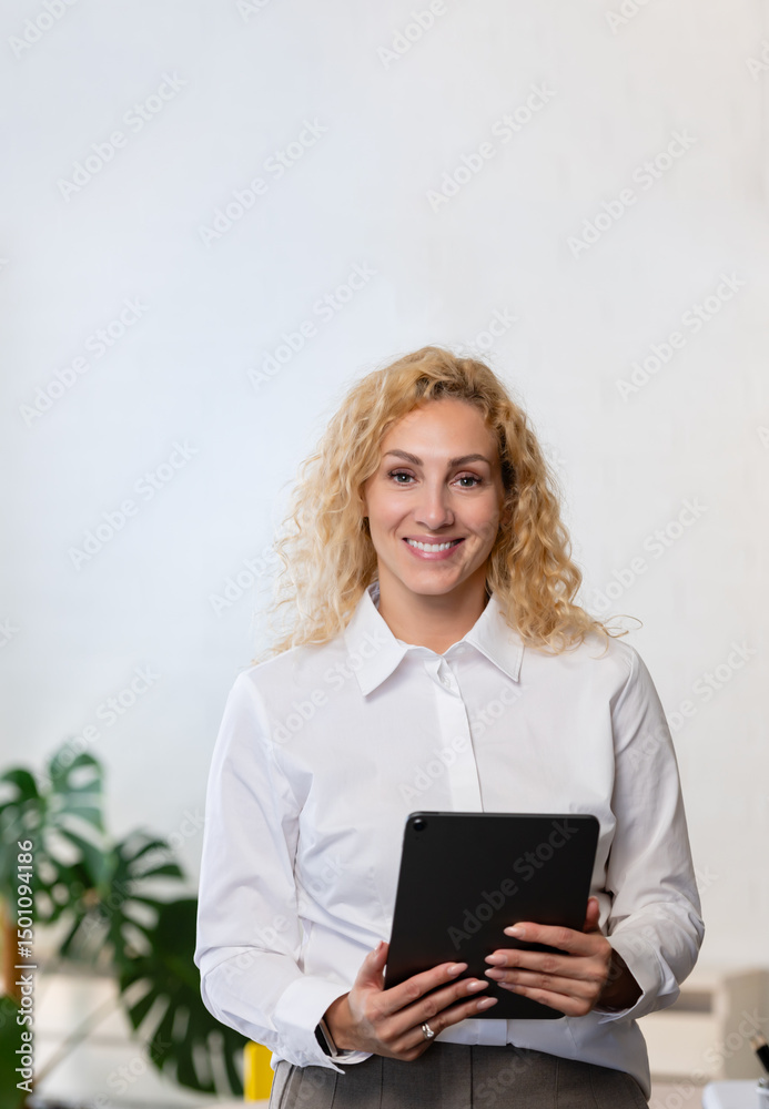 Fototapeta premium Smiling businesswoman holding a tablet in a bright modern office. Confident and approachable, she stands in a professional environment with plants and minimal decor.