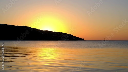 Tranquil seascape at sunset with golden reflections on the calm water and the sun setting behind a silhouette of a coastal hill.