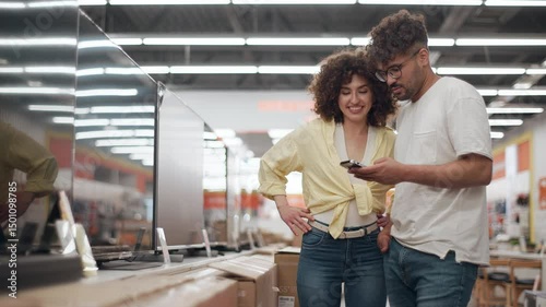 Customers choosing television in electronics store. Young couple browsing television options, comparing devices and sharing joyful moment while selecting modern electronics in sleek retail environment
