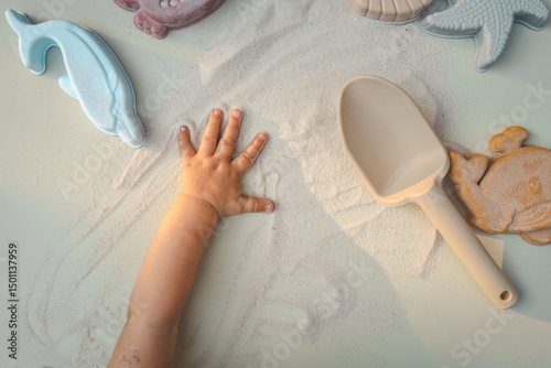 Wallpaper Mural Top view of a child’s hand playing with sand, surrounded by pastel-colored sea creature toys and a beige scoop. The hand leaves playful marks in the sand, evoking sensory exploration and summer joy Torontodigital.ca