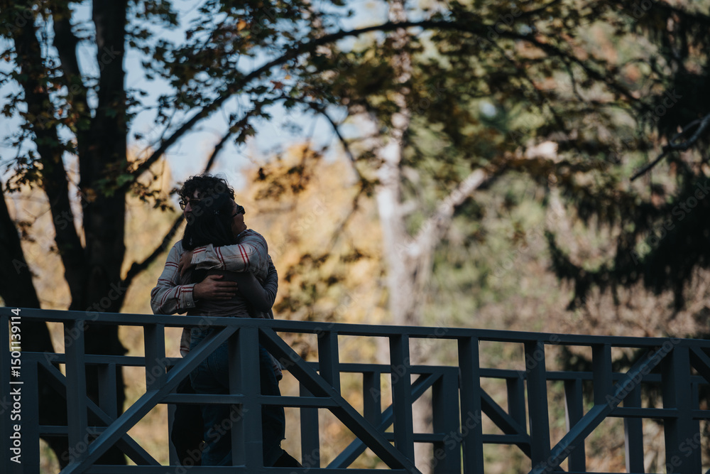 Obraz premium A loving couple embraces on a wooden bridge amidst a lush forest, capturing a moment of affection and tranquility. Sunlight filters through the trees, creating a romantic atmosphere.