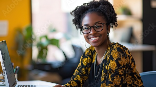 A smiling woman with braided hair is seated at a desk, wearing an orange button-up shirt.