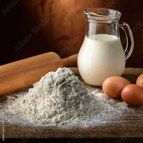 A pile of flour with fresh eggs, a jug of milk and a rolling pin with a rustic background