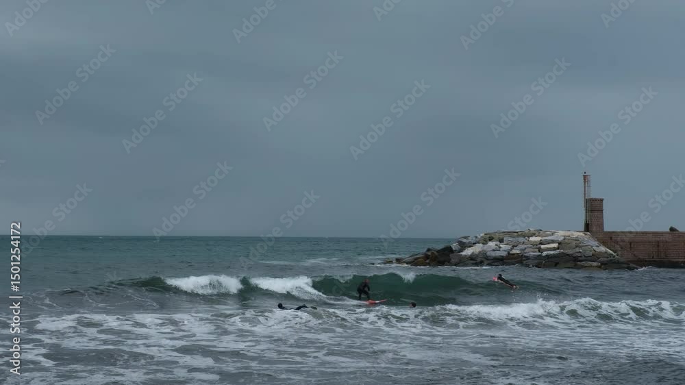 Recco, Italy - 17.04.2025: Surfers in the sea trying to ride waves. Athletes in wetsuits surf small waves on an overcast day with a lighthouse in the background. Recco, Italy - 17.04.2025: Surfers in the sea trying to ride waves. Athletes in wetsuits surf small waves on an overcast day with a lighthouse in the background.