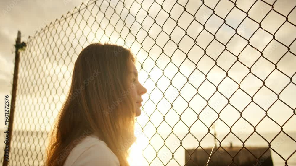 Young woman observing sunset through metal mesh fence