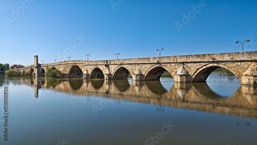 Panoramic image of the historic Old Bridge over the Maritsa River in Svilengrad, Bulgaria, reflecting beautifully in the calm blue water under a clear sky.