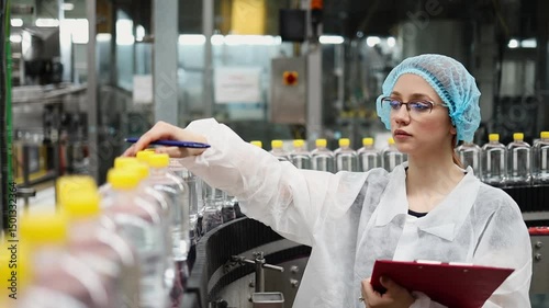 Female quality control inspector wearing protective clothing and mask writing notes on clipboard while inspecting production line in water bottling factory