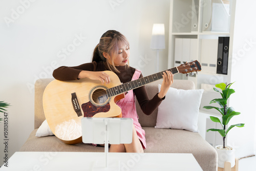 Beautiful young Asian woman with acoustic guitar sitting on sofa and concentrating on learning to play using app on tablet placed nearby
