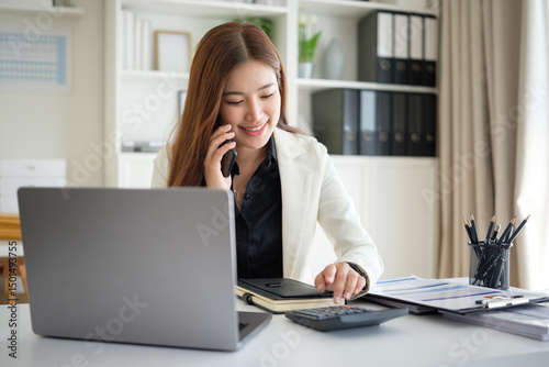 Professional woman in office attire talking on the phone while working on financial tasks with a calculator and documents at her desk.