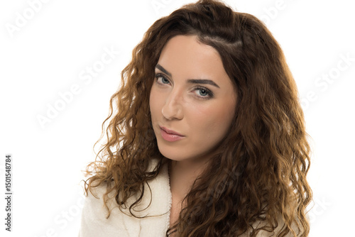 Woman with curly brown hair in white bathrobe with suspicious expression against white background, skeptical look, raised eyebrow, natural portrait
