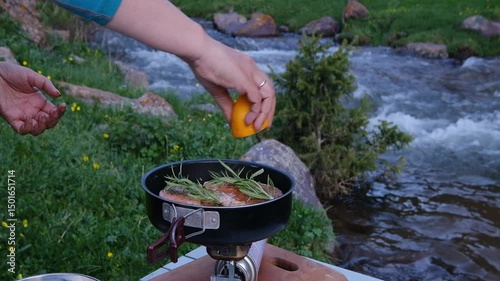 Close-up of female hands squeezing fresh lemon on salmon steaks with rosemary sprigs in a frying pan over a portable camping gas stove. Cooking outdoors by a mountain river, surrounded by wildflowers 