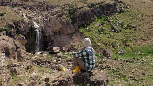 A female traveler in a checkered shirt, knitted hat and yellow boots sits on a rock opposite a small steppe waterfall. Nature of Kazakhstan waterfall in the Malaysary ridge in the Almaty region.