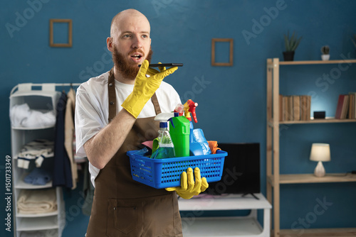 Young bearded man wearing gloves and apron holding basket with cleaning supplies dictate voice message using phone. Male maid in domestic duties. Household, home cleaning and housework