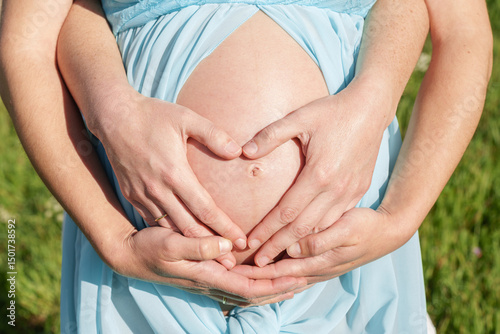 Close-up detail of a pregnant woman with her dress open, holding her belly and being held by her partner's hands.