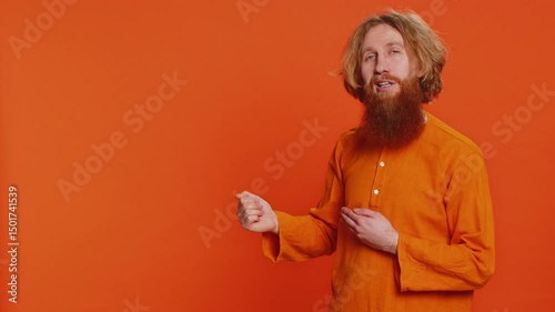 Caucasian bearded man showing thumbs up and pointing left empty place, advertising area for commercial text copy-space for goods promotion advertisement. Guy isolated on orange background. Lifestyles