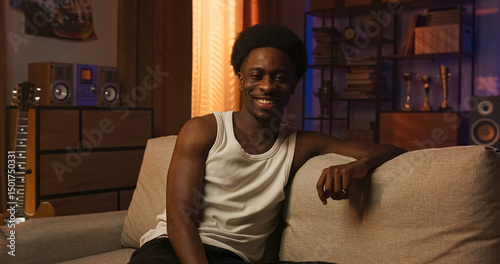 A happy young man of multi-racial background sits on a couch in warm natural light, gazing confidently at the camera and laughing with genuine joy and inner peace.