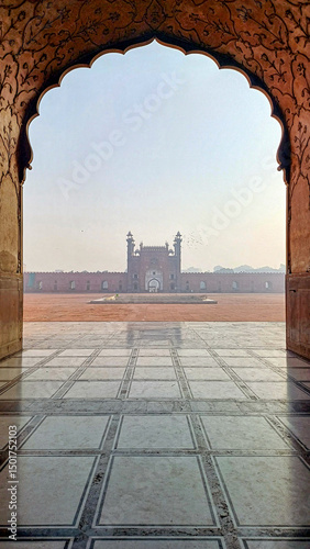 An ornate archway, adorned with Mughal floral patterns, frames a hazy view of the Badshahi Mosque's red sandstone facade and courtyard. The foreground shows a patterned marble floor