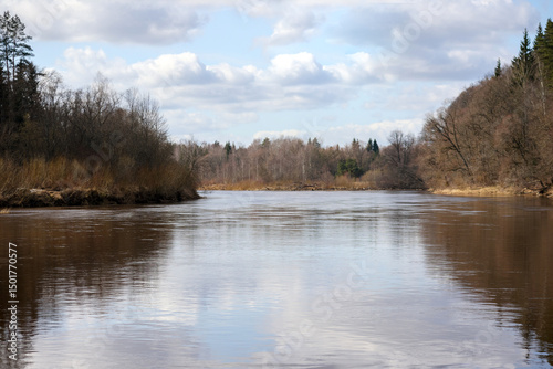 
a wide river surrounded by trees and shrubs with bare leaves, its water reflecting the sky and the surroundings.