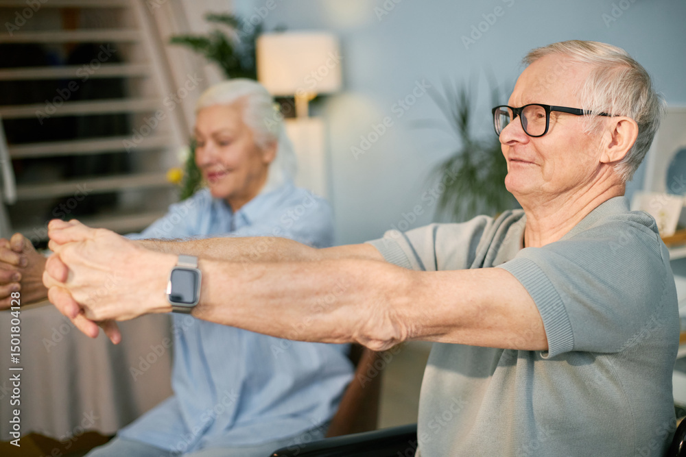 Fototapeta premium Senior Caucasian man performing stretching exercises with elderly woman smiling in the background, showcasing an active lifestyle in a cozy living room environment