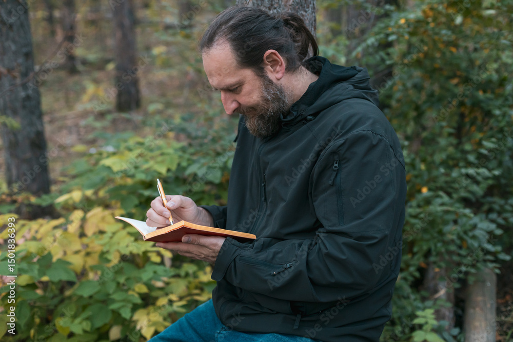 Fototapeta premium A man with a beard is sitting in a forest and writing in a notebook