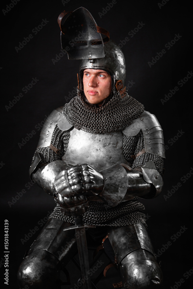 Naklejka premium Portrait of a knight in full plate armor holding a sword posing sitting on chair isolated on a dark background.