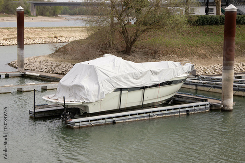 Boat covered with a protective tarp to shield from sun and rain, parked near a pier in a European marina, showing careful storage and weather protection.