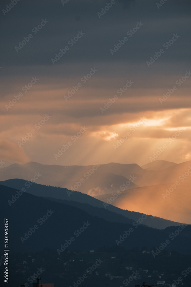 Fototapeta premium Mountain layers under a dramatic sunset sky Beams of light pierce through clouds illuminating the peaks