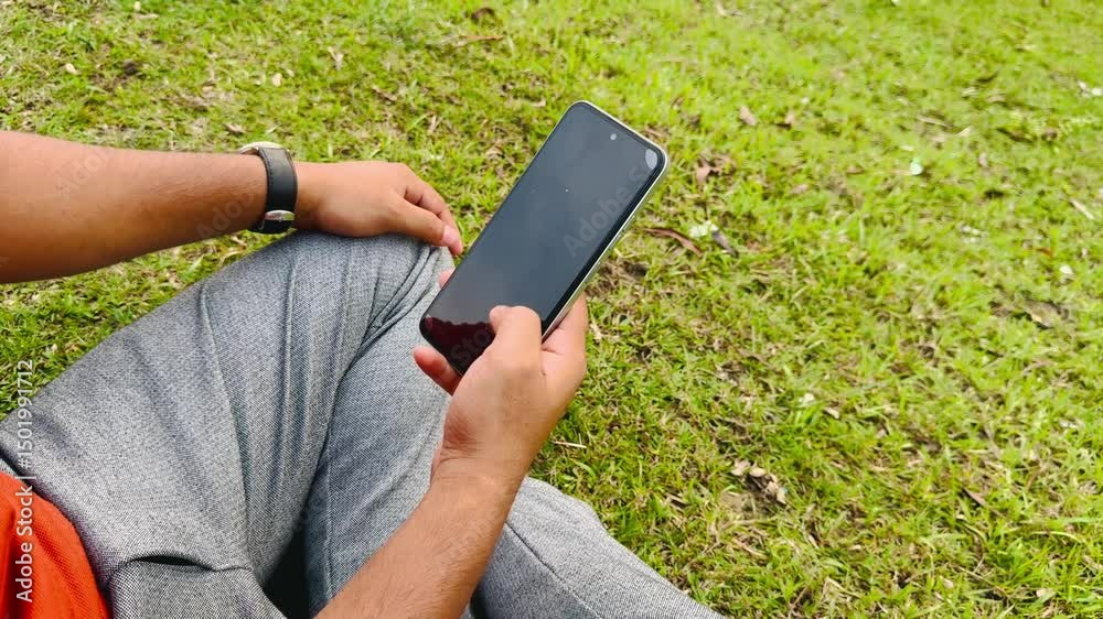 Man Using Smartphone While Sitting on Green Grass in Park, Typing and Reading Messages

