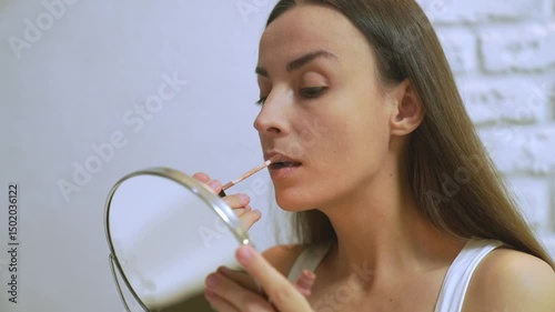Young woman applying nude lipstick while looking into a handheld mirror. She is wearing a white tank top, standing against a white brick wall in soft indoor lighting.
