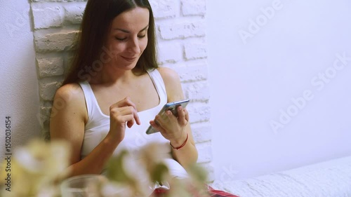 A young woman in a white tank top sits indoors by a white brick wall, smiling and scrolling on her smartphone, creating a relaxed and modern home lifestyle atmosphere.
