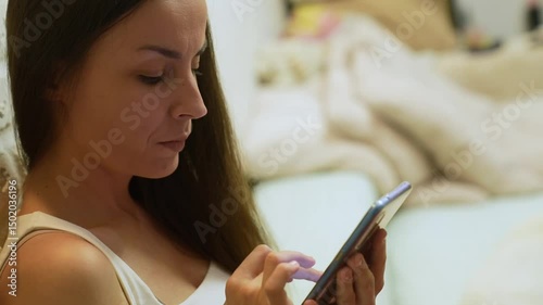 Young woman in a white tank top sitting indoors on a bed, focused on using her smartphone. She scrolls or types with concentration in a cozy, softly lit setting.
