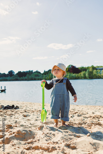 Toddler in a panama hat on his head and with bare feet walks on the sand on the beach. 2 year old boy digs golden sand on a warm sunny day. Child on a walk on the beach.