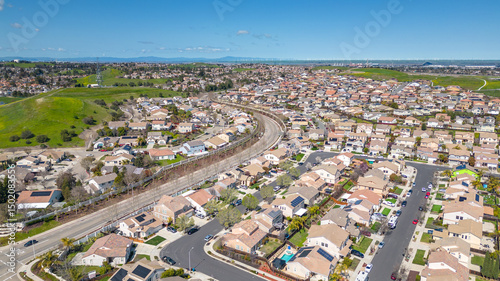 Aerial view of a suburban neighborhood and shopping center in Antioch, California, on a clear spring day with scenic hills, green fields, and residential homes with solar panels.