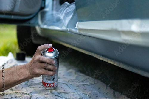 Male hand applying paint to lower car body with a spray can in a DIY repair session. Real auto maintenance in action