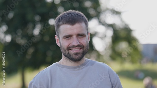 A man with a cheerful expression stands in a park, surrounded by greenery, enjoying a sunny day while exuding a sense of calm and happiness.