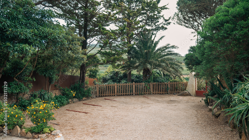 Obraz premium Secluded gravel parking area in a remote countryside location, surrounded by lush tropical vegetation, tall trees, and a wooden fence, creating a peaceful and natural setting