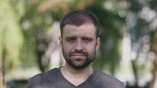 Man stands in a park, wearing a casual outfit and expressing a friendly demeanor. The setting features bright sunlight filtering through nearby trees.