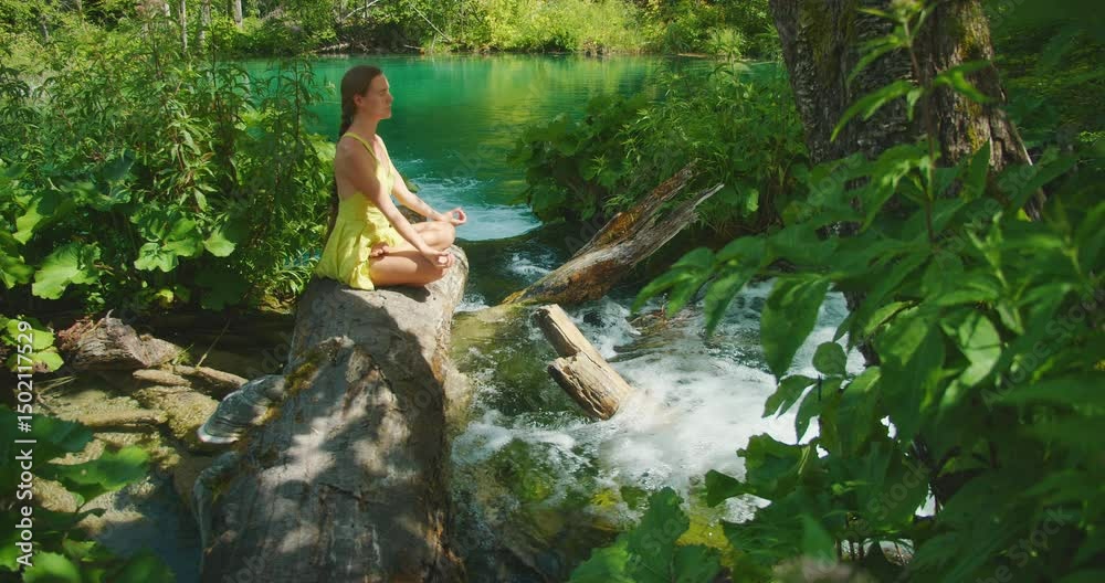 Woman meditates on a log above running water in serene summer forest