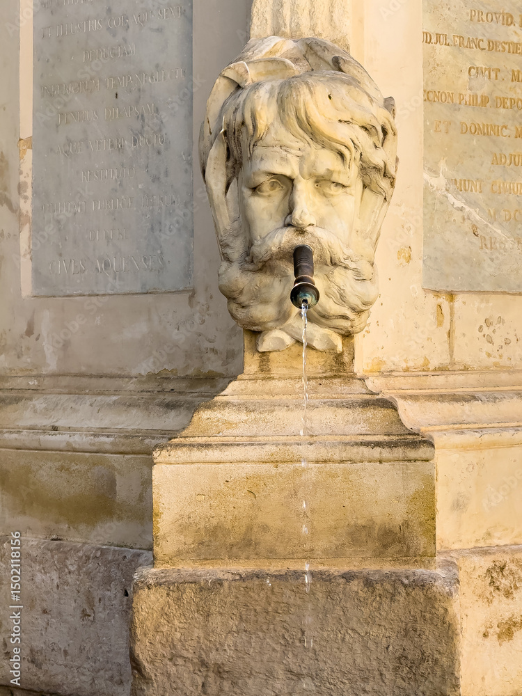 Obraz premium Aix-en-Provence, France - July 10, 2024: Fontaine de l'eau benite, holy water, on Place de l'hotel de ville. Detail showing water flowing out of the mouth of the male head sculpture