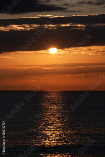 Sunset over the ocean, vertical background image. Setting sun appearing from behind clouds.