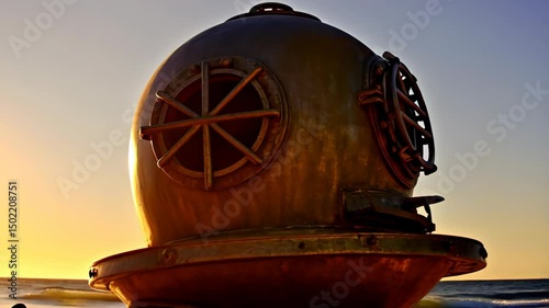 Antique diving helmet standing on a sandy beach during sunset reflecting in the waves creating a nostalgic seaside scene