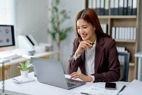 Asian businesswoman using laptop and holding pen while sitting at desk in office, working on project, analyzing financial reports, browsing internet, smiling