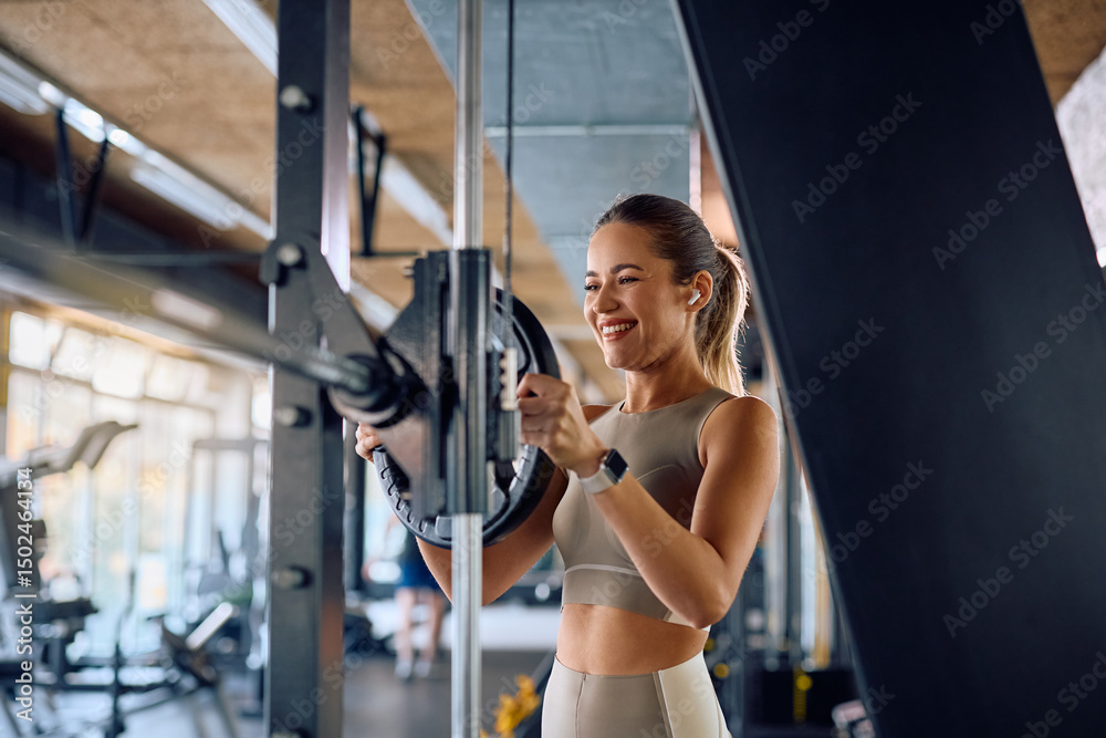 Fototapeta premium Happy sportswoman adjusting weight disk on barbell during strength training in gym.