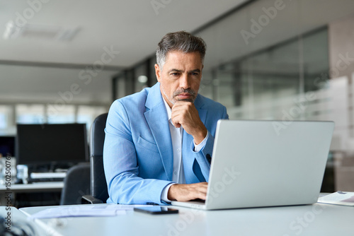 Busy 50 year old senior businessman executive, serious middle aged professional business man financial investor wearing suit using laptop computer working on investment project sitting at office desk.