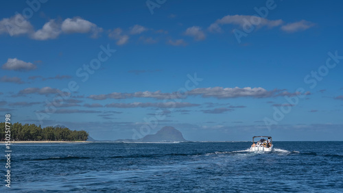 A tourist motorboat is sailing on the blue ocean. Foam trail behind the stern. Ripples on the surface of the water. Green vegetation on the coast of the island. Mountain  against azure sky and clouds.