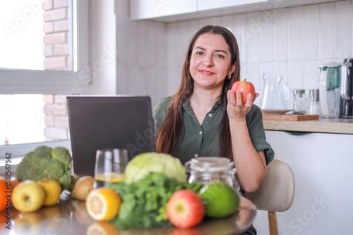 woman holding apple in hand, sitting in kitchen with laptop and preparing meal plan, among fresh vegetables and fruits. home cooking concept, vegetarianism