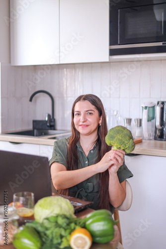 woman holding broccoli, researching healthy recipes on laptop among fresh products in kitchen. food blogging concept, diets, healthy eating