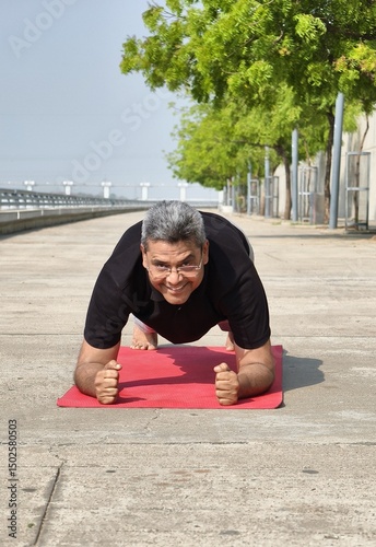 Senior Indian man doing forearm plank on a yoga mat outdoors, smiling and enjoying his fitness routine in the fresh morning air at Riverfront, Ahmedabad. Man is staying fit after 60 years of age. 